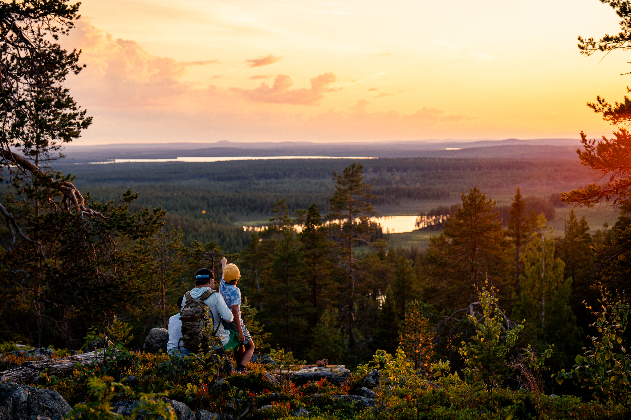 Isä ja kaksi lasta istuvat vaaran laidalla ja ihailevat kesäistä auringonlaskua. Kaukana näkyy vesistöjä ja metsää ilta-auringon loisteessa.
